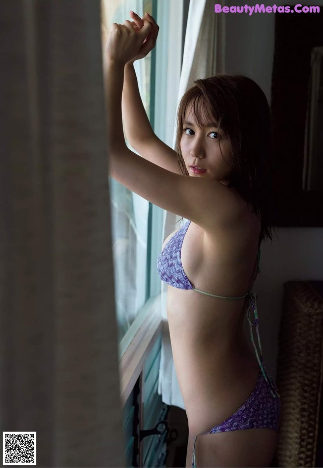 A woman in a purple bikini standing by a window.