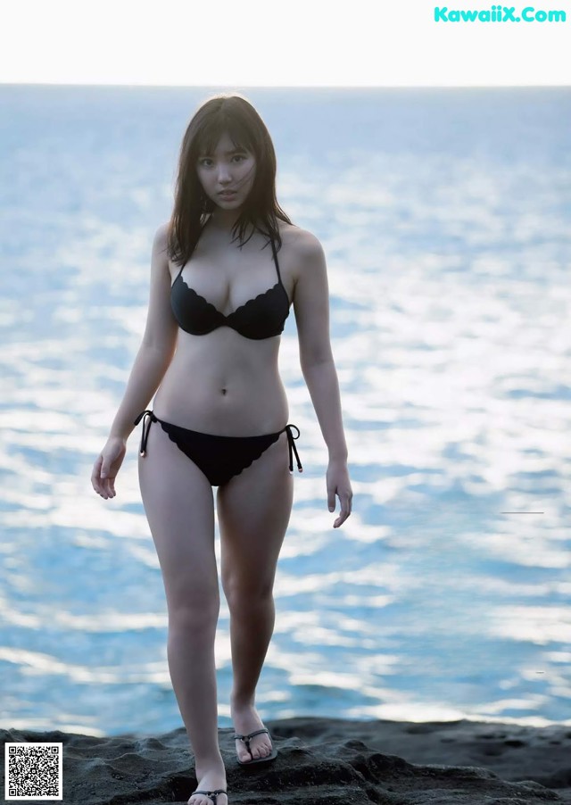 A woman in a black bikini standing on a rock by the ocean.