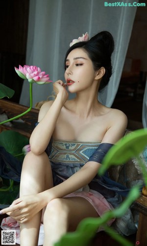 A woman in a blue dress sitting on a bed next to a pink flower.